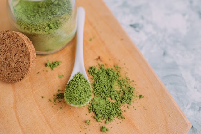 Kratom powder in a white gram spoon next to a jar of kratom powder