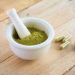 white kratom in a small white bowl with kratom capsules laying on a wooden table top next to the bowl