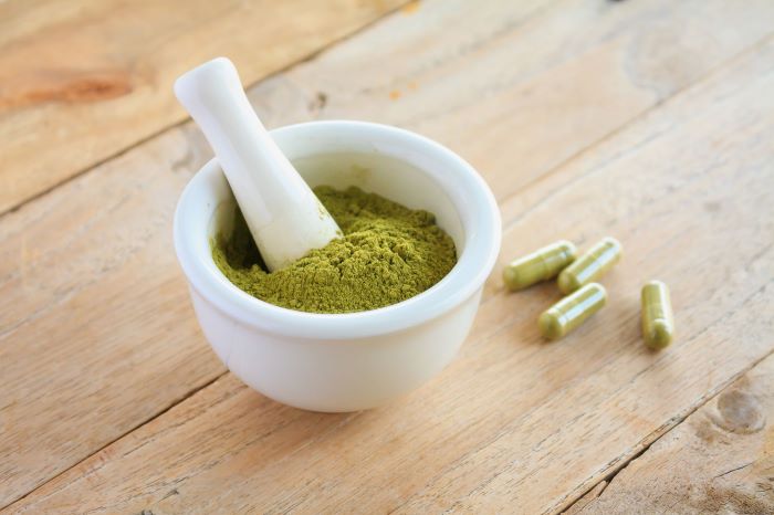 white kratom in a small white bowl with kratom capsules laying on a wooden table top next to the bowl